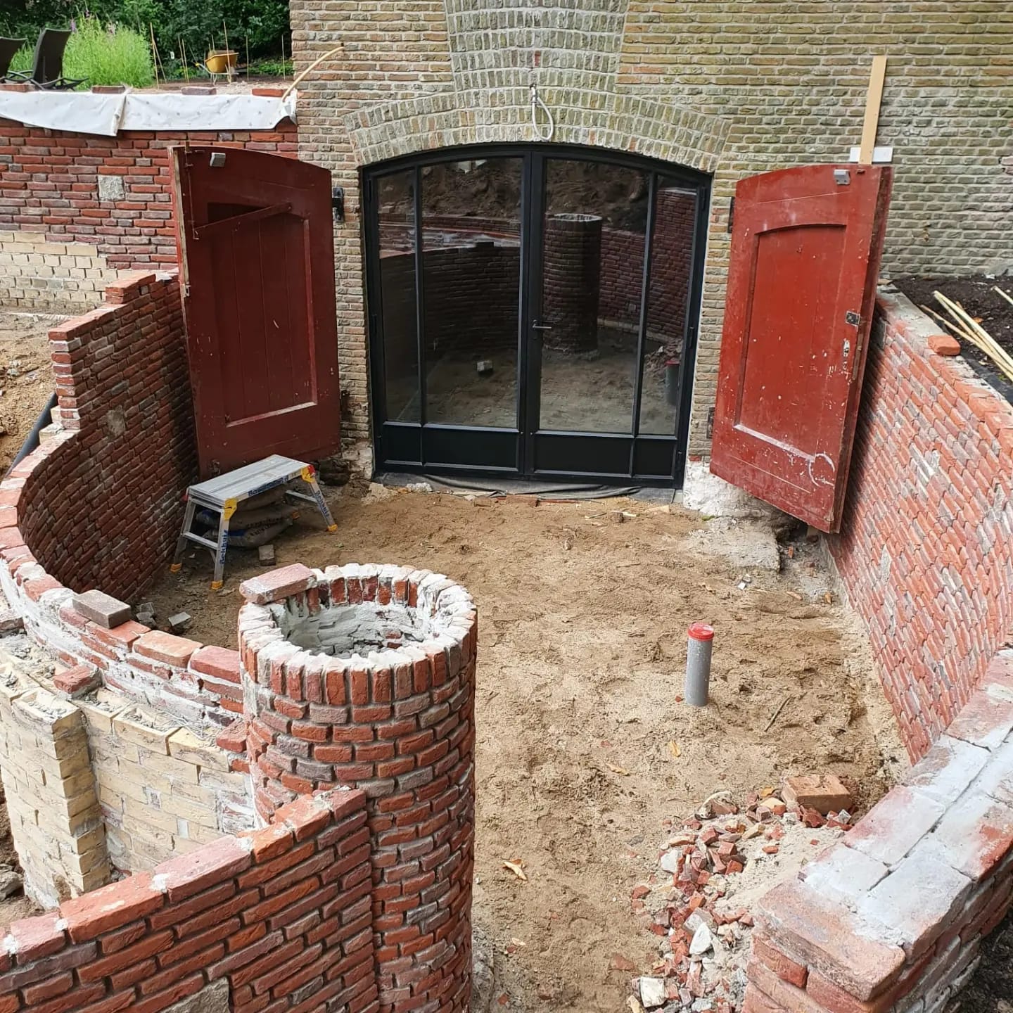 Gebogen gemetselde tuinmuur met een gemetselde put, rode houten deuren en stalen boogdeuren in een oude bakstenen gevel